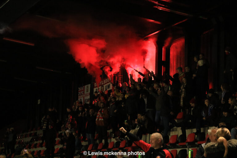 Fans of FC United of Manchester light red torches on the stands as they welcome their team to the pitch