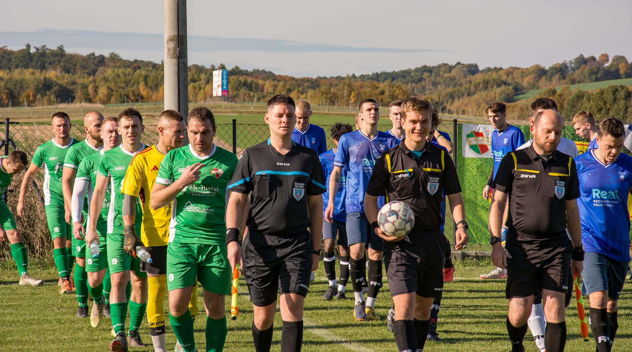 Victoria Smroków and Krakow Dragoons FC players, together with referees, entering the field