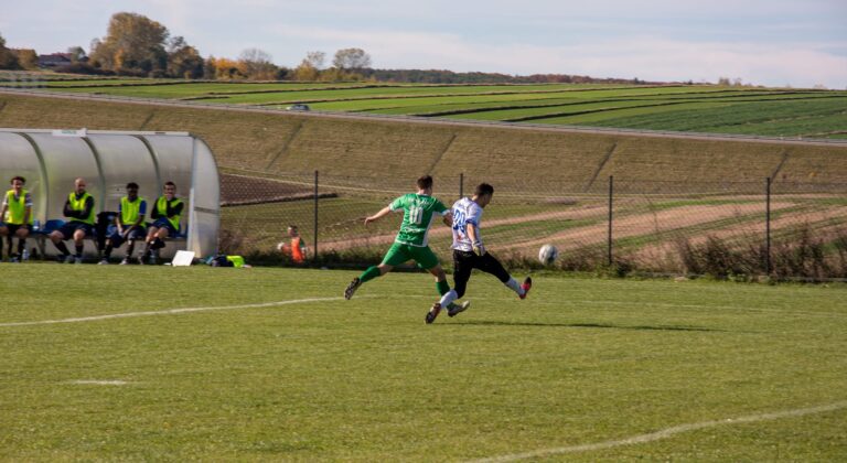 Marcin Obyrtacz of Krakow Dragoons FC kicks a ball out of danger. Countryside fields visible in the background.